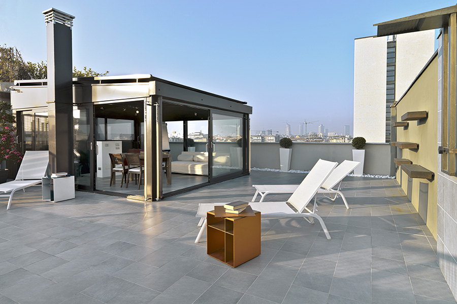 Blick auf stylische Dachterrasse mit modernem, hellgrauem Fliesenböden. Auf der Terrasse stehen weiße Liegestühle und ein Wintergarten mit Couch und Esstisch ist angeschlossen.